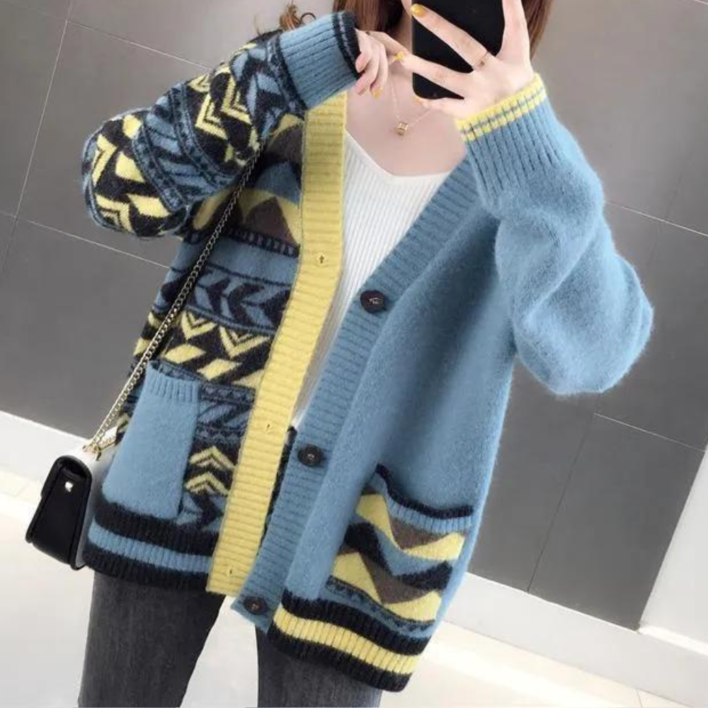 Vrouw draagt blauwe gebreide cardigan met geometrisch patroon en gele accenten, gecombineerd met witte top en zwarte tas. Modieuze dameskleding.