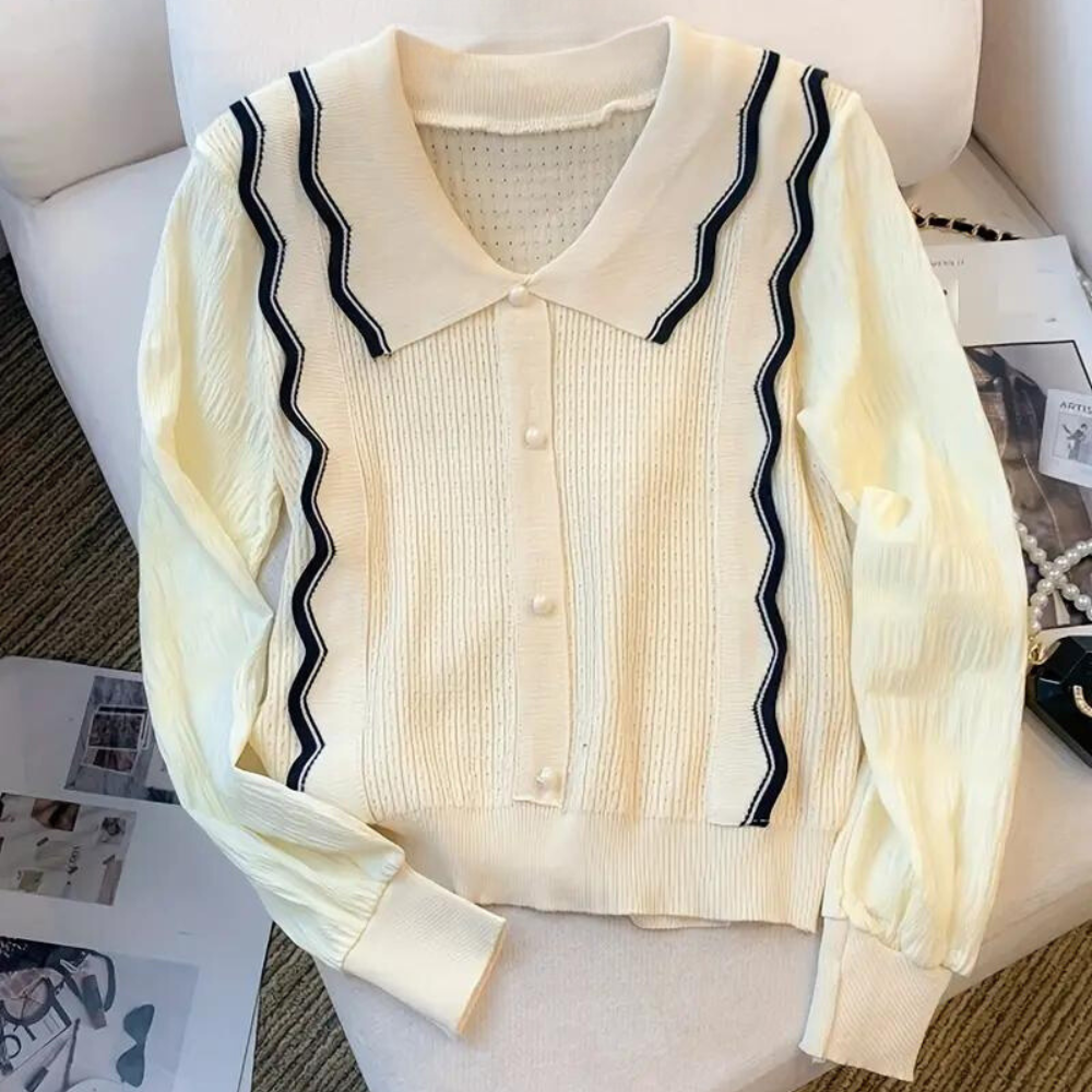 Crèmekleurig damesvest met zwarte zigzagstrepen, knoopsluiting en kraag, liggend op een witte bank. Stijlvol en trendy kledingstuk.