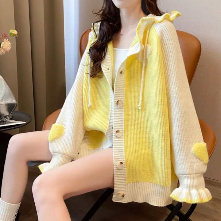 Vrouw draagt gele en witte gebreide cardigan met knopen en capuchon, zittend op stoel. Modieuze dameskleding, herfst/winter stijl.