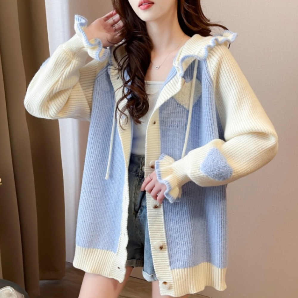 Vrouw draagt een gebreid vest in blauw en crème met hartpatroon, gecombineerd met een wit topje en denim shorts. Modieuze dameskleding, casual stijl.