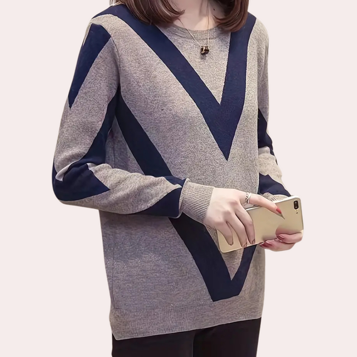 Vrouw in grijze trui met donkerblauwe V-strepen, houdt smartphone vast. Modieuze dameskleding, casual stijl, herfst-winter mode.