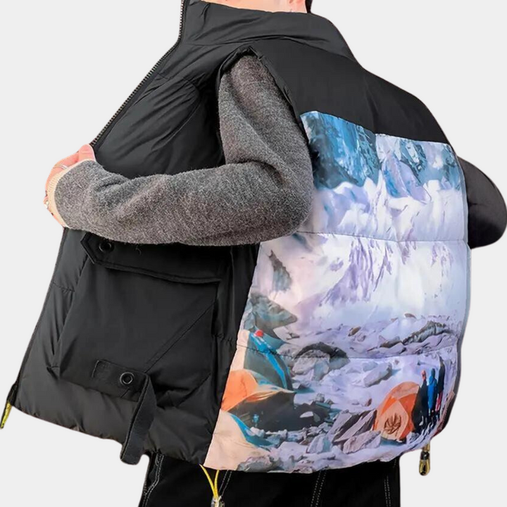 Zwarte gewatteerde bodywarmer met kleurrijke berglandschapprint op de rug, gedragen door persoon in grijze trui. Modieuze outdoor kleding.