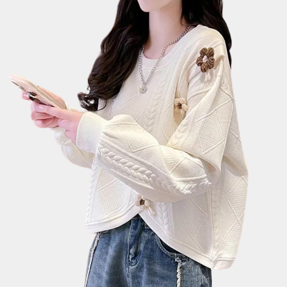 Vrouw in crèmekleurige trui met kabelpatroon en bloemenaccenten, draagt spijkerbroek. Modieuze dameskleding, herfsttrui, casual outfit.