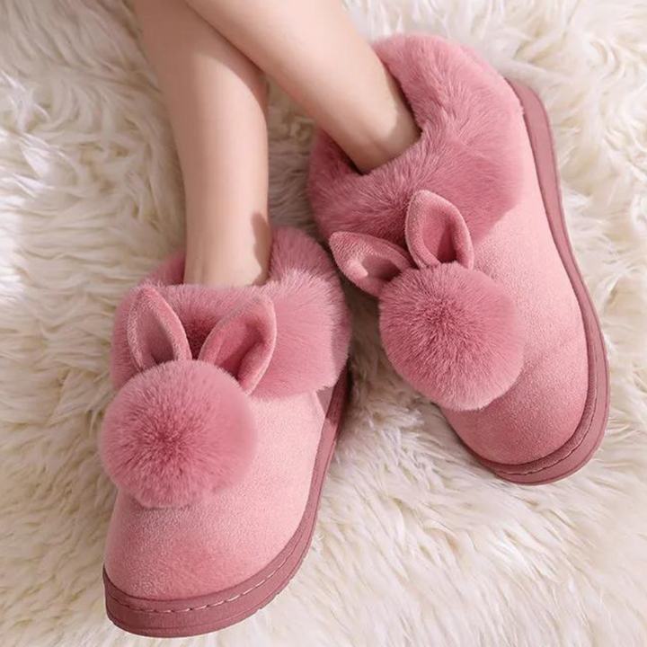 Roze pluche pantoffels met konijnenoren en pompon, gedragen door een persoon op een witte, zachte ondergrond. Warme winterpantoffels voor dames.