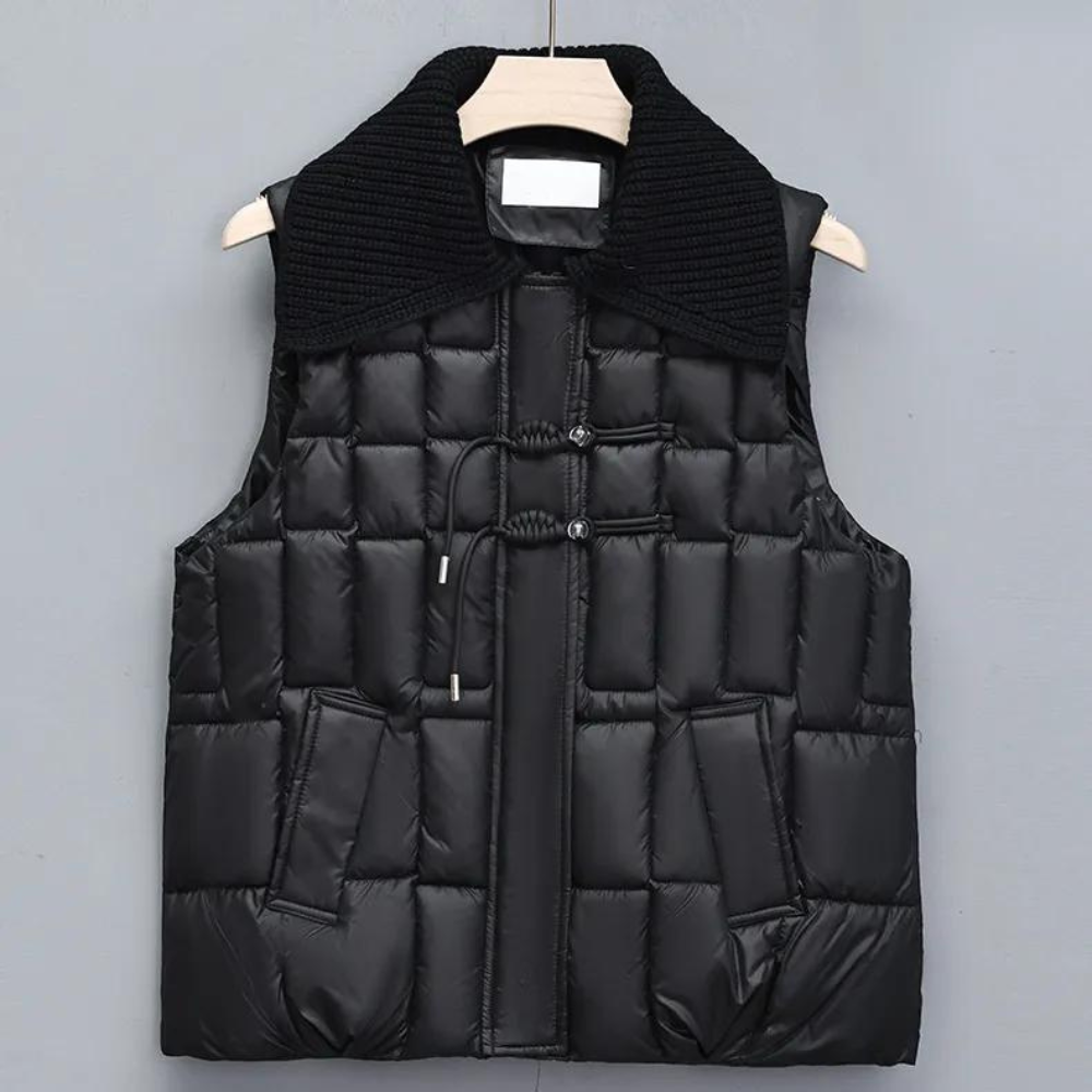 Zwarte gewatteerde bodywarmer met brede kraag, knoopsluiting en zakken, hangend aan een houten hanger tegen een grijze achtergrond.