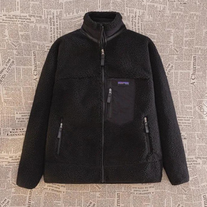 Zwarte fleece jas met rits, borstzak en twee zijzakken, hangend voor een achtergrond met krantenprint. Warme winterkleding, unisex design.