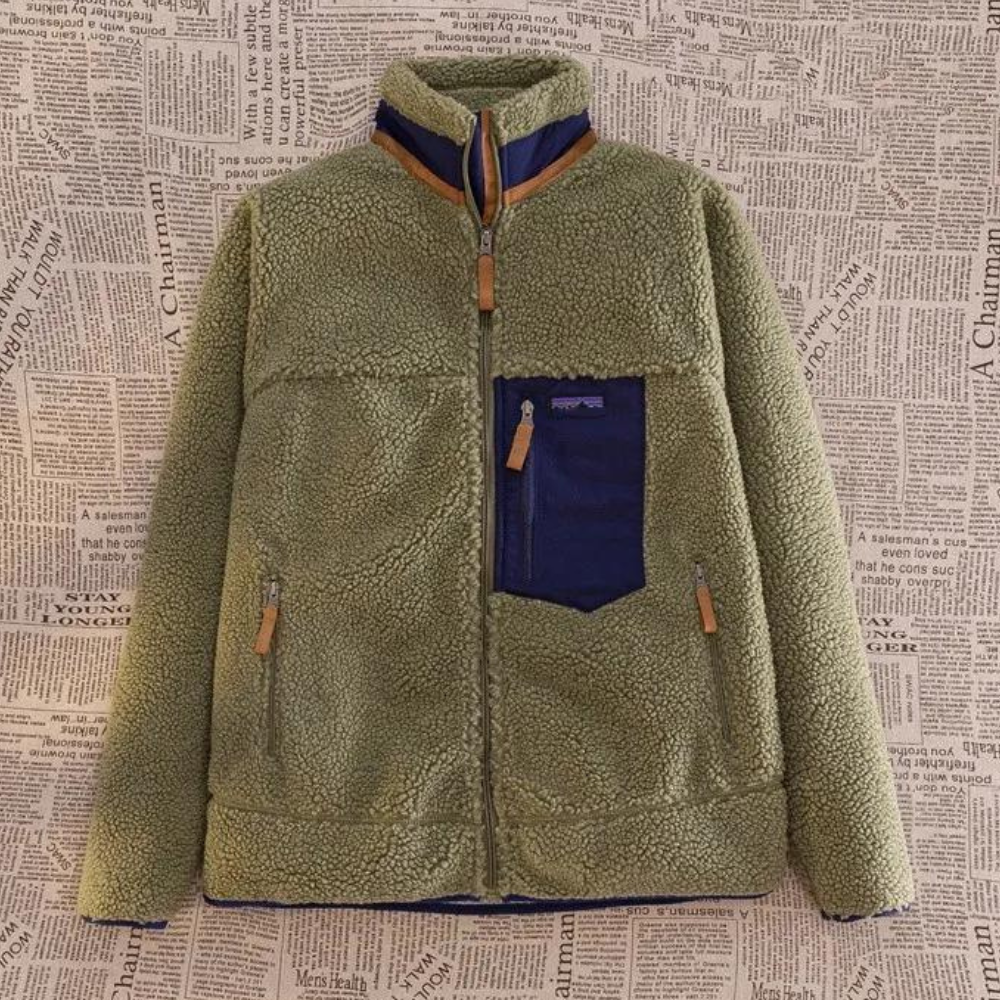 Groene fleece jas met rits en blauwe borstzak, hangend voor een achtergrond met krantenprint. Warme winterkleding, modieuze outdoorstijl.