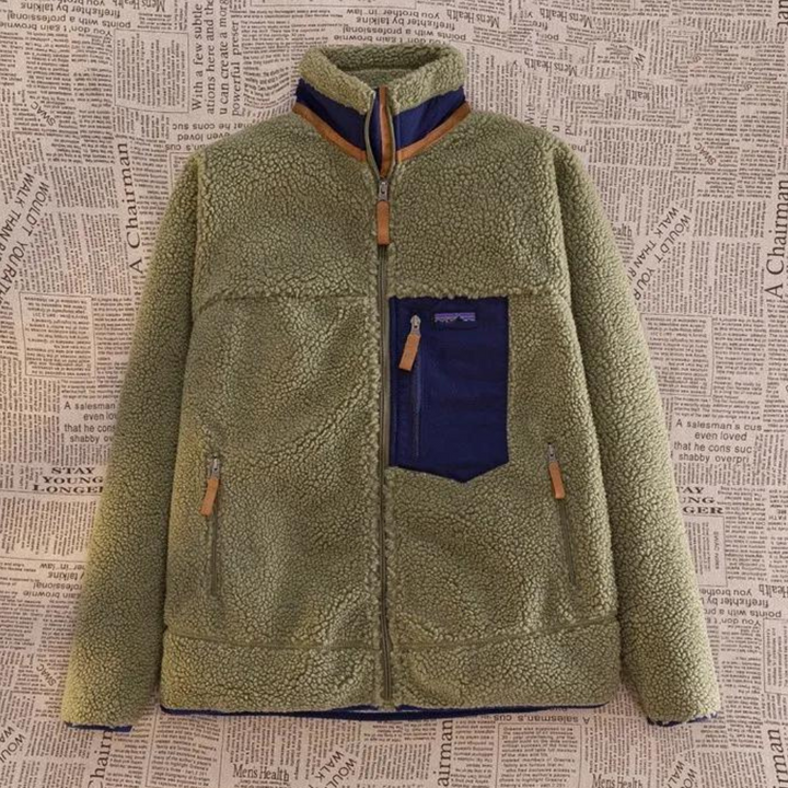 Groene fleece jas met rits en blauwe borstzak, hangend voor een achtergrond met krantenprint. Warme winterkleding, modieuze outdoorstijl.