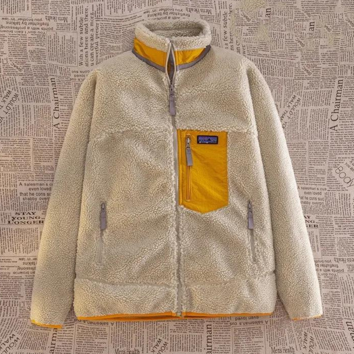 Beige fleecejack met gele accenten, ritsen en borstzak, hangend voor een achtergrond van krantenprint. Warme winterkleding, stijlvol en functioneel.