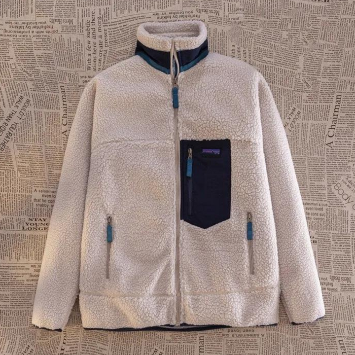Witte fleece jas met blauwe accenten, ritsen en borstzak, tegen een achtergrond van krantenprint. Warme winterkleding, stijlvol en functioneel.