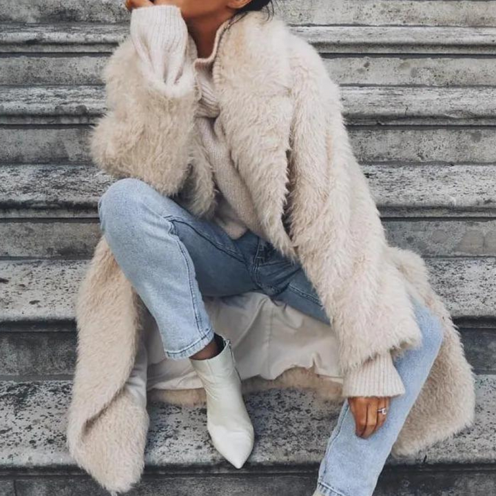 Vrouw in beige teddyjas, blauwe jeans en witte laarzen zittend op stenen trap. Modieuze winteroutfit, trendy dameskleding, streetstyle.