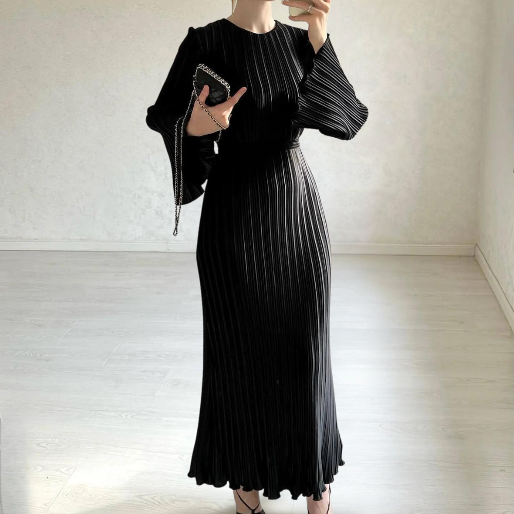 Elegante zwarte geplooide jurk met lange mouwen, gedragen door een vrouw in minimalistische kamer. Perfect voor avondkleding en modieuze evenementen.