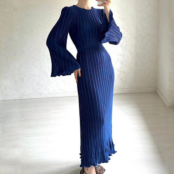 Vrouw in elegante, lange blauwe plissé jurk met wijde mouwen, staand in minimalistische kamer. Perfect voor avondkleding en modieuze evenementen.