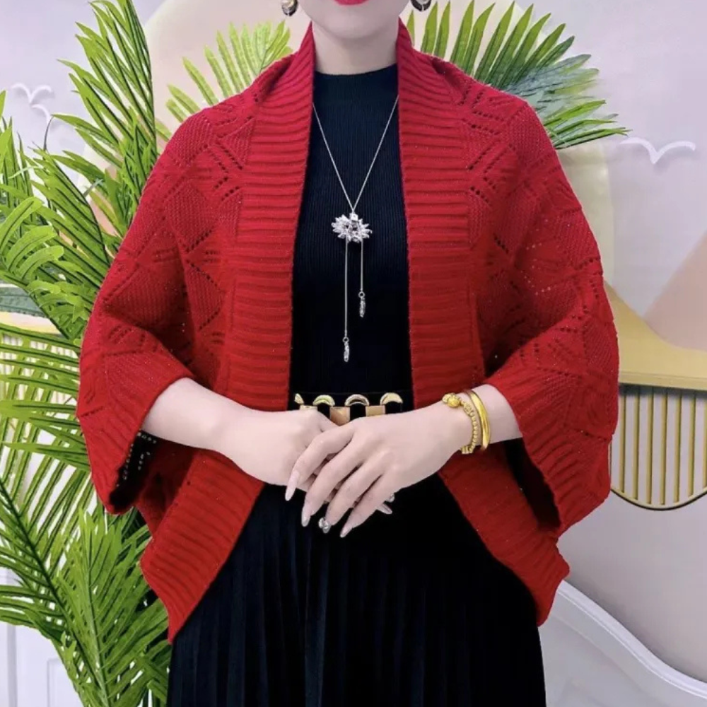 Vrouw in rode gebreide omslagdoek met zwart jurkje, gouden sieraden en planten op achtergrond. Mode, herfstkleding, stijlvolle damesoutfit.