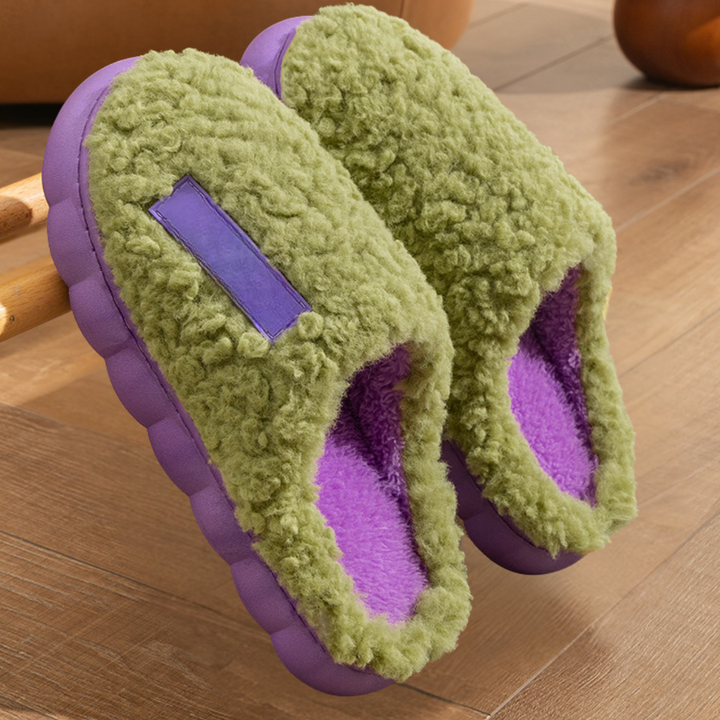 Groene pluche pantoffels met paarse zool en binnenvoering, liggend op houten vloer. Comfortabele huisschoenen, ideaal voor warme voeten thuis.