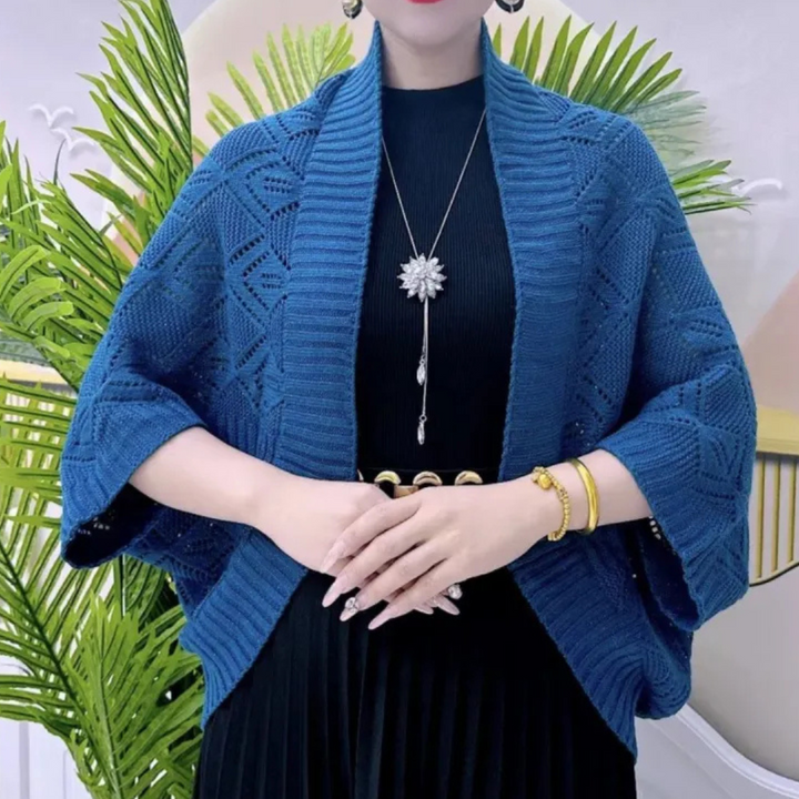 Vrouw draagt blauwe gebreide omslagdoek met geometrisch patroon, zwarte jurk en opvallende ketting. Stijlvolle damesmode, herfstkleding.