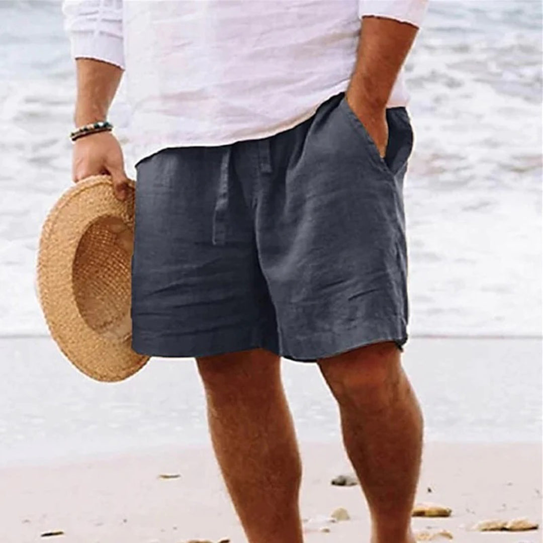 Man in witte blouse en donkergrijze linnen korte broek op strand, houdt strohoed vast. Zomerse strandmode, casual herenkleding, comfortabele stijl.