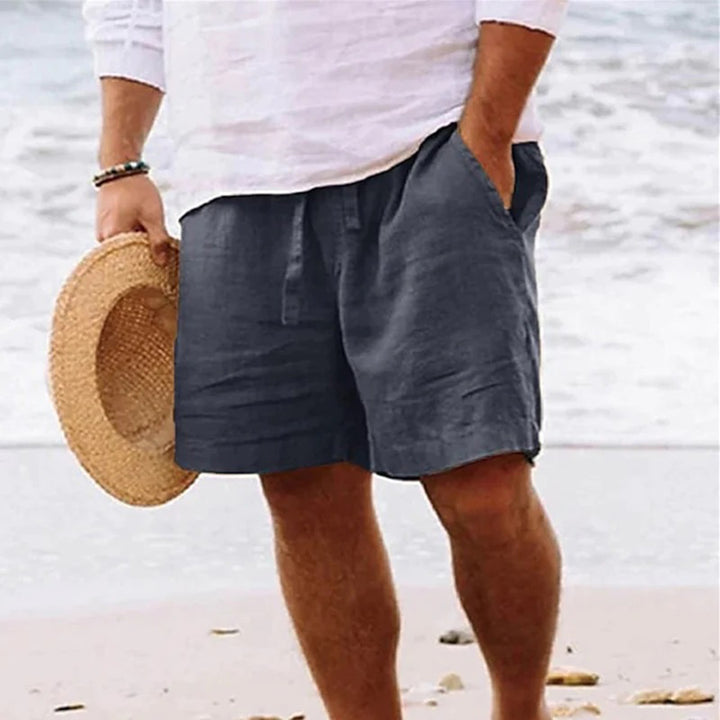 Man in witte blouse en donkergrijze linnen korte broek op strand, houdt strohoed vast. Zomerse strandmode, casual herenkleding, comfortabele stijl.