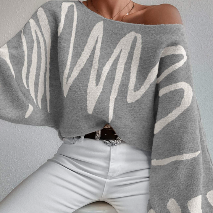 Grijze oversized trui met abstracte witte zigzagpatronen, off-shoulder ontwerp, gedragen door model met witte broek. Trendy damesmode, herfstkleding.