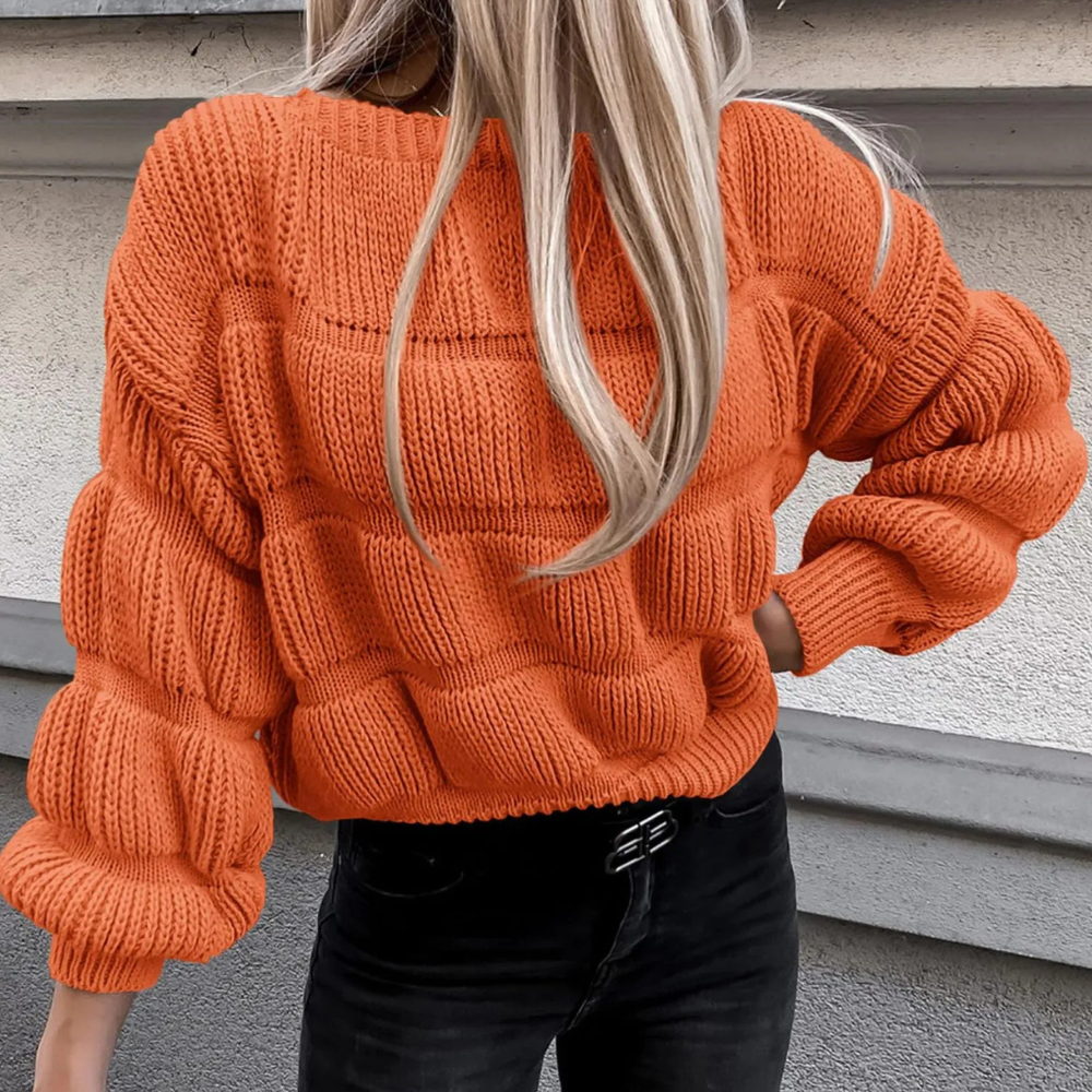 Oranje gebreide trui met blokpatroon, gedragen door vrouw met lang blond haar, gecombineerd met zwarte jeans. Trendy herfstmode, casual stijl.
