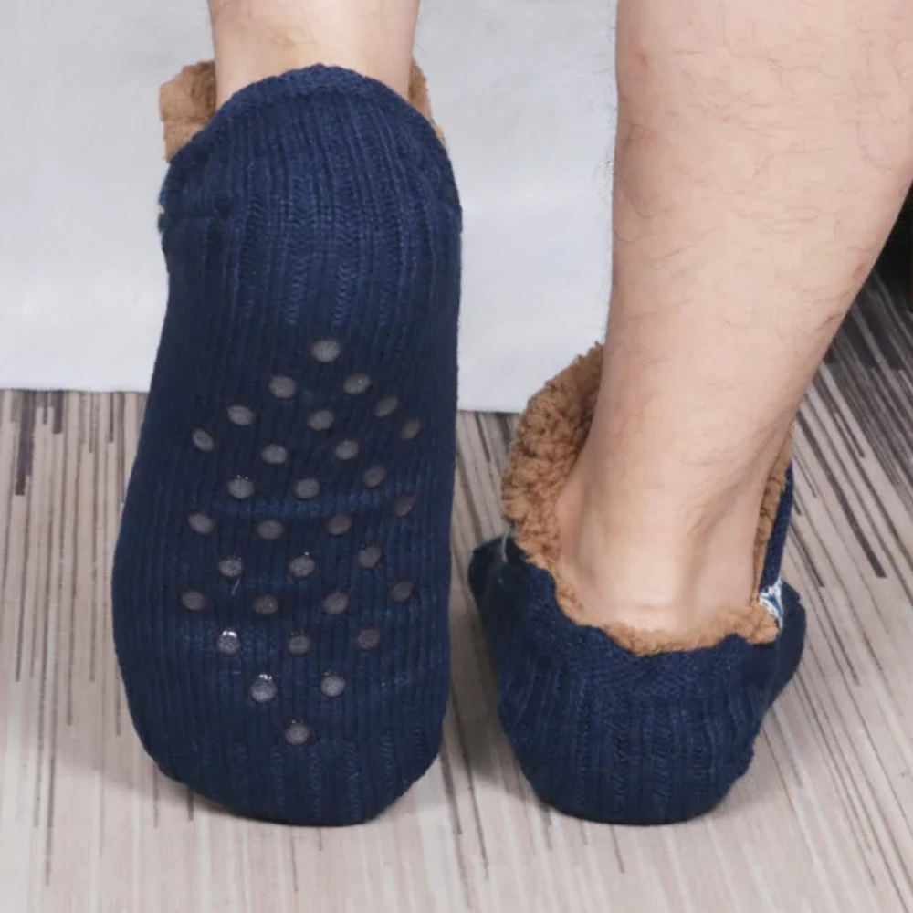 Blauwe antislip huissokken met bruine voering, gedragen door een persoon op een houten vloer. Comfortabele warme sokken voor thuisgebruik.