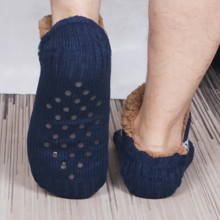 Blauwe antislip huissokken met bruine voering, gedragen door een persoon op een houten vloer. Comfortabele warme sokken voor thuisgebruik.