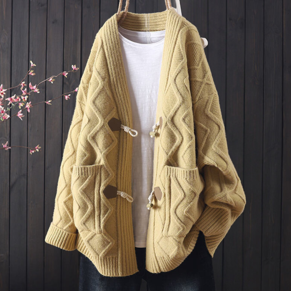 Beige gebreid vest met kabelpatroon, houten knopen en zakken, hangend voor een donkere houten achtergrond. Comfortabele herfstmode.