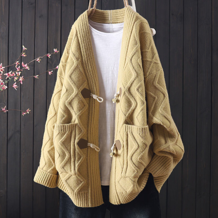 Beige gebreid vest met kabelpatroon, houten knopen en zakken, hangend voor een donkere houten achtergrond. Comfortabele herfstmode.