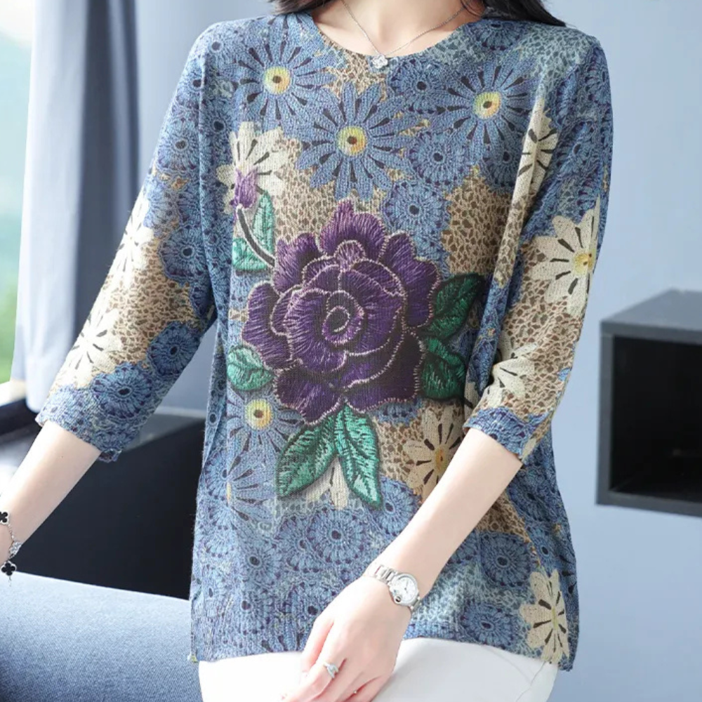 Vrouw draagt een blauwe blouse met kleurrijke bloemenprint, waaronder een grote paarse roos. Modieuze dameskleding, casual stijl, lente outfit.