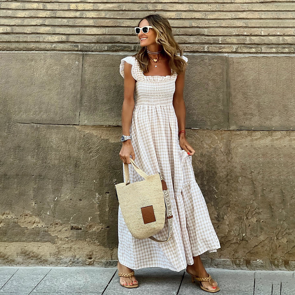 Vrouw in lange zomerjurk met ruitpatroon, draagt zonnebril en strooien tas, poseert tegen stenen muur. Perfecte outfit voor zomerse dagen.