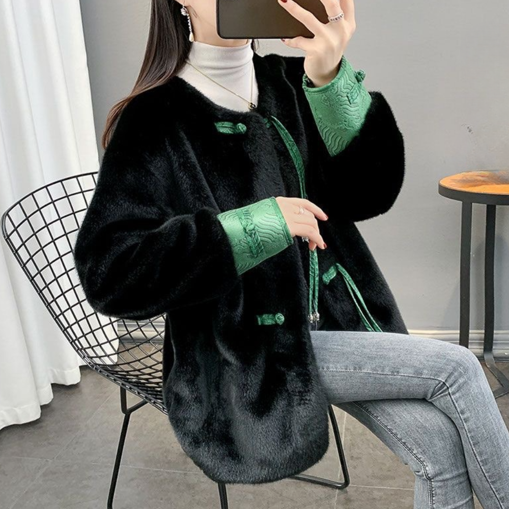 Vrouw in zwarte faux fur jas met groene accenten, zittend op metalen stoel, draagt grijze jeans en witte coltrui. Wintermode, stijlvolle outfit.