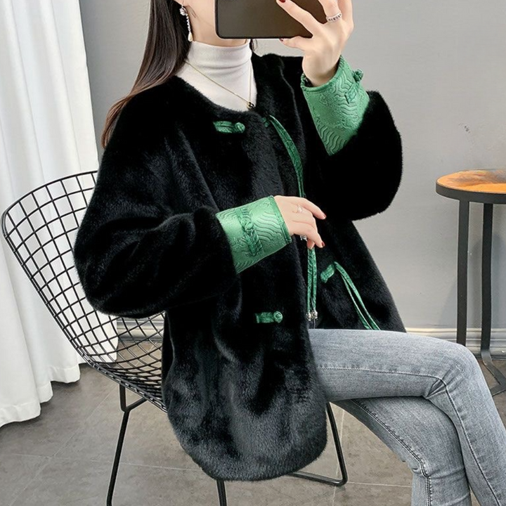 Vrouw in zwarte faux fur jas met groene accenten, zittend op metalen stoel, draagt grijze jeans en witte coltrui. Wintermode, stijlvolle outfit.