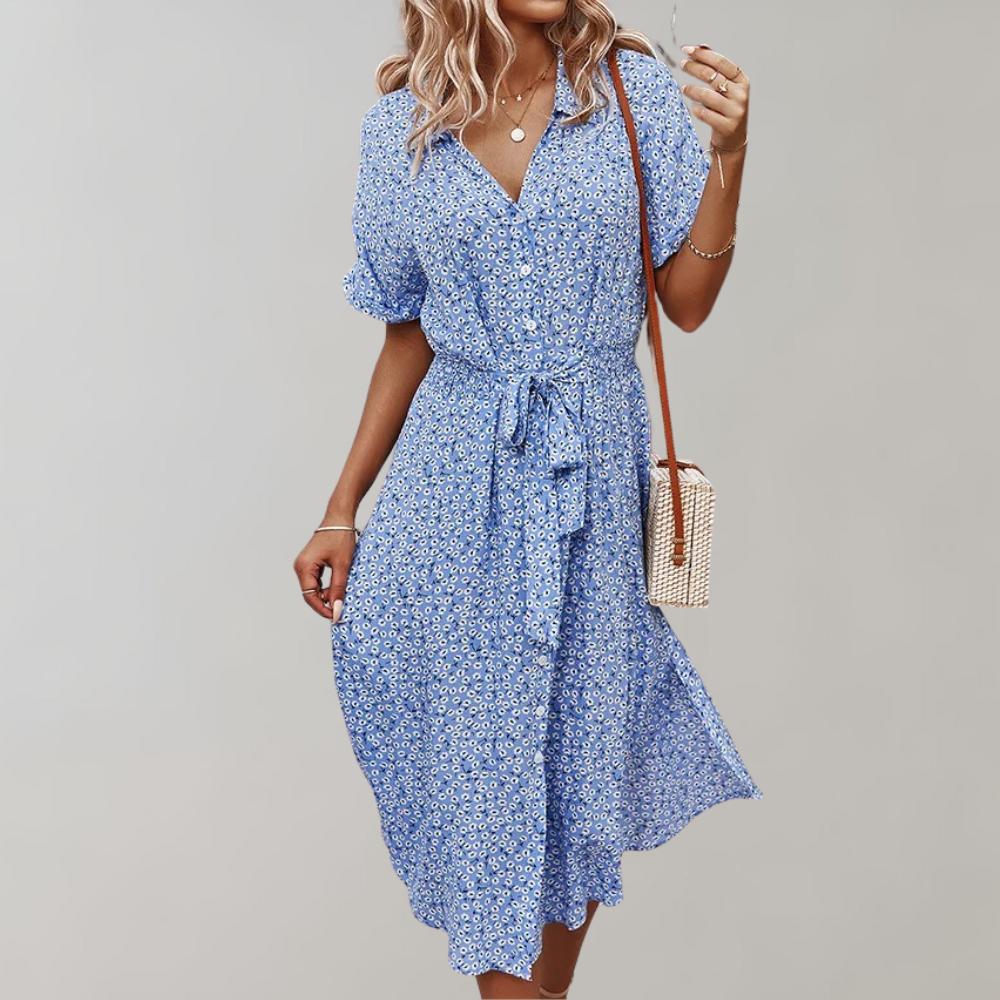 Vrouw in blauwe midizomerjurk met bloemenprint, korte mouwen en strikceintuur, draagt een schoudertas. Perfecte damesmode voor warme dagen.