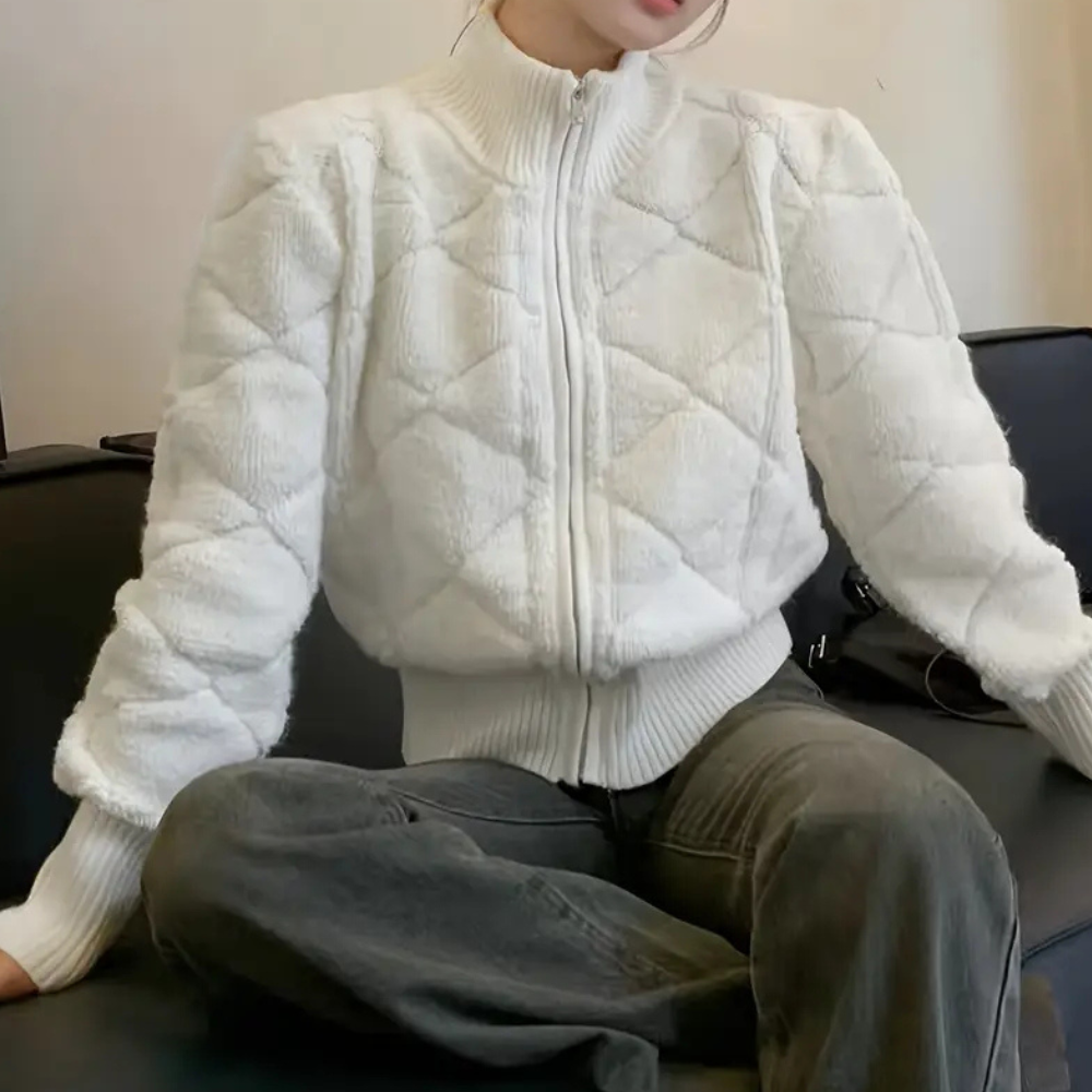 Vrouw draagt witte gewatteerde jas met ruitpatroon, zittend op zwarte bank. Modieuze winterkleding, trendy damesjas, comfortabele outfit.