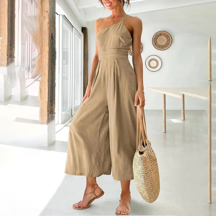 Vrouw in stijlvolle kaki jumpsuit met asymmetrische schouder, draagt een gevlochten tas en sandalen in moderne, minimalistische woonkamer.