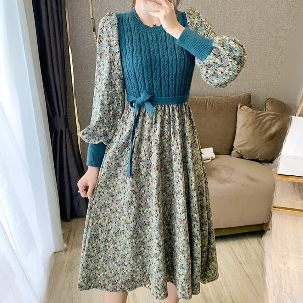 Vrouw in bloemenjurk met blauwe gebreide top en strik, staand in moderne woonkamer. Elegante damesmode, herfstkleding, trendy outfit.