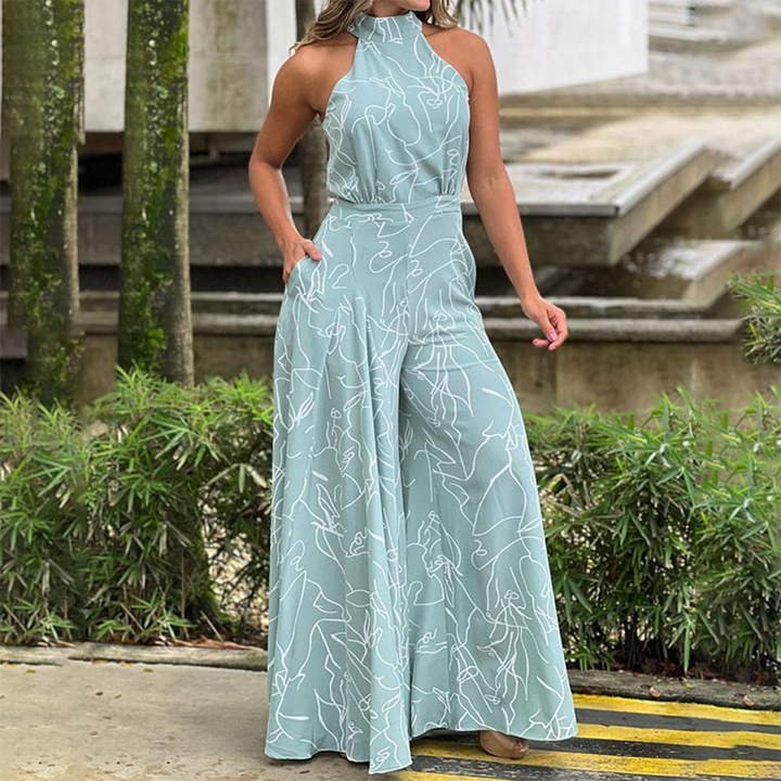Vrouw in lichtblauwe jumpsuit met abstracte witte lijnen, staand buiten bij groene planten. Modieuze dameskleding, zomeroutfit.