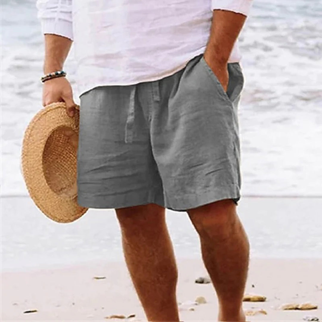 Man in lichtgrijze linnen korte broek en wit overhemd, houdt strohoed vast op strand. Zomerse herenkleding, casual strandoutfit.