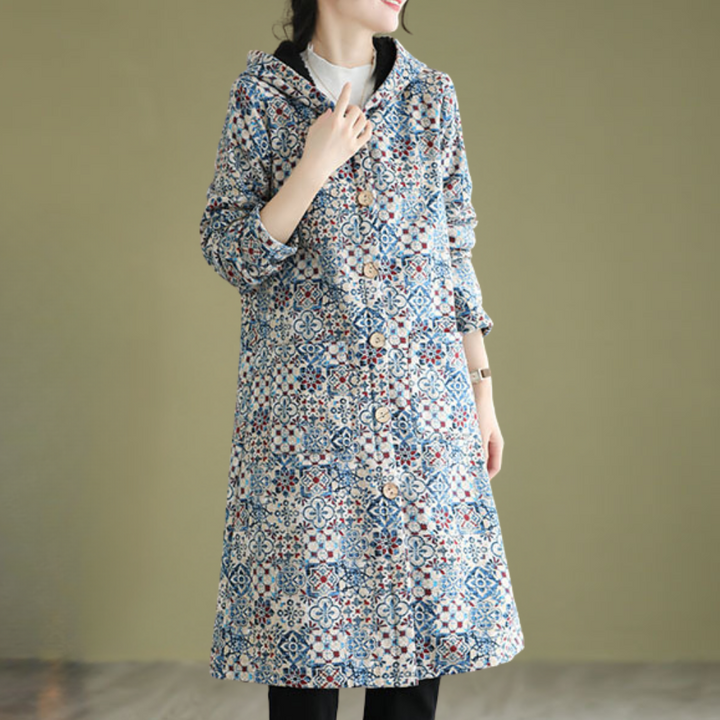 Vrouw draagt lange, kleurrijke jas met geometrisch patroon en knopen, tegen een neutrale achtergrond. Modieuze dameskleding, herfst/winter stijl.