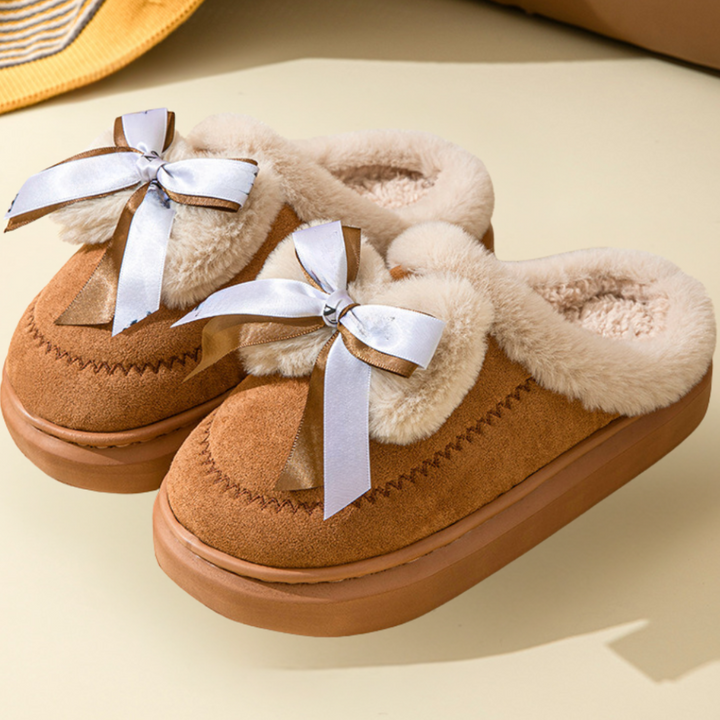Bruine damespantoffels met witte strik, pluche voering en verhoogde zool. Perfect voor wintercomfort en stijl. Warme, modieuze huisschoenen.