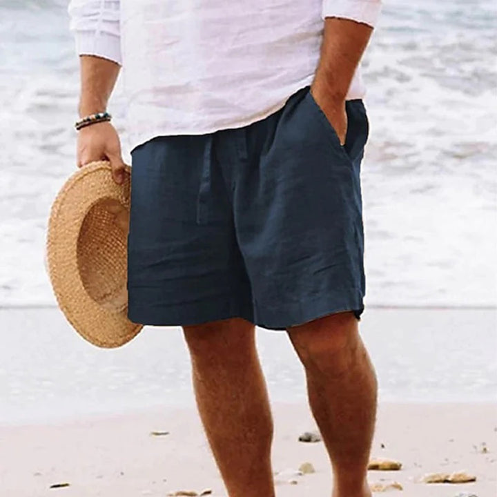 Man in witte blouse en marineblauwe korte broek op strand, houdt strohoed vast. Zomerse strandmode, casual herenkleding, ontspannen stijl.