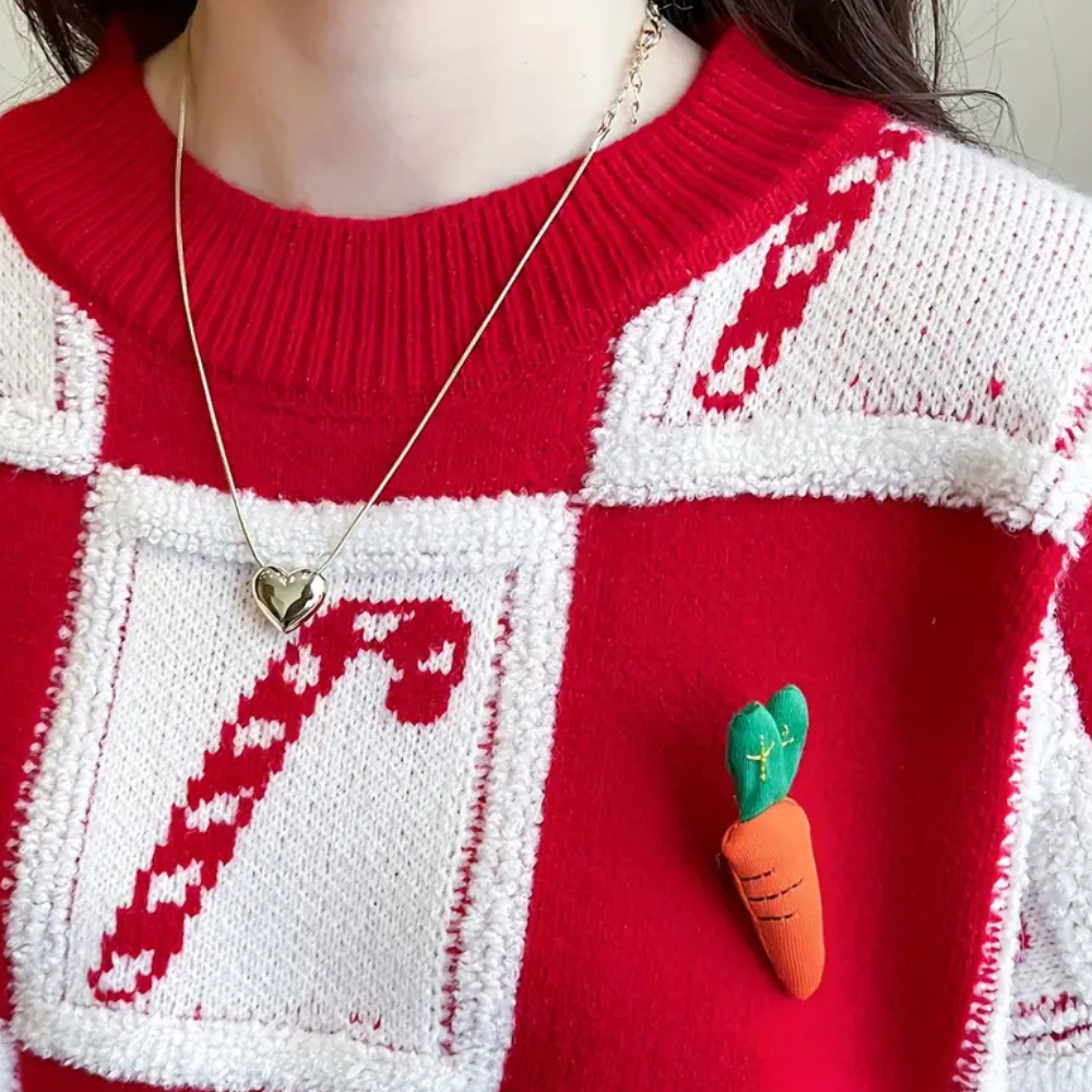 Rode kersttrui met witte vierkanten, snoepstokpatroon en wortelbroche, gedragen door persoon met hartvormige ketting. Perfect voor feestdagen.