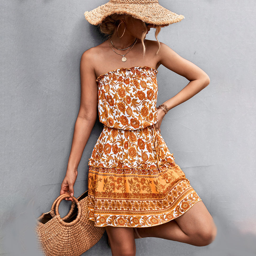 Vrouw in zomerse oranje bloemenjurk met strohoed en rieten tas, tegen grijze achtergrond. Perfecte outfit voor strandmode en zomerse stijl.