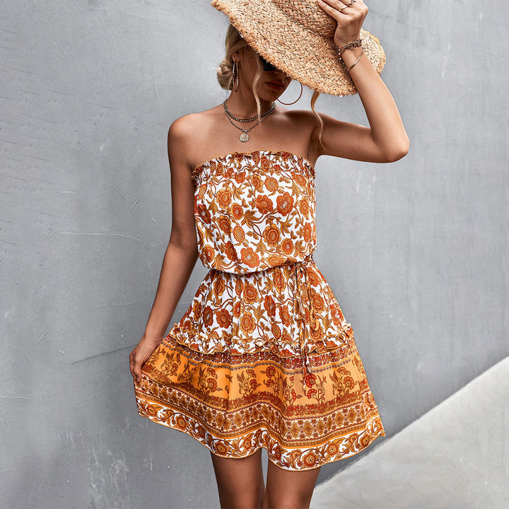Vrouw in oranje zomerjurk met bloemenprint en strohoed, poseert tegen een grijze muur. Boho-chic mode, casual zomeroutfit, strandkleding.