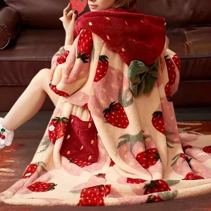 Vrouw in een warme, aardbeienprint fleece deken met capuchon, zittend op een bruine bank. Perfect voor knusse winteravonden thuis.