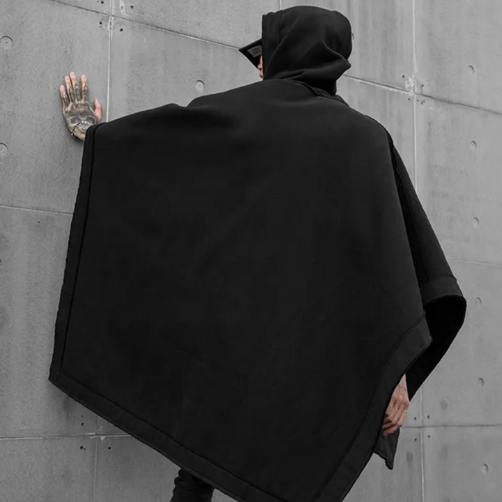 Zwarte unisex poncho met capuchon, gedragen door persoon tegen betonnen muur. Streetwear mode, minimalistisch design, urban stijl.
