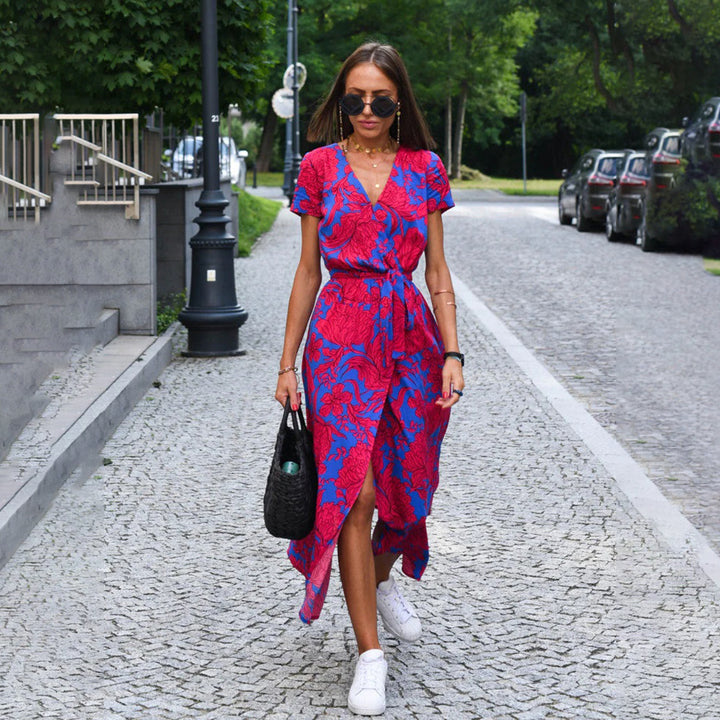 Vrouw in kleurrijke rood-blauwe jurk loopt op geplaveide straat, draagt zwarte tas en witte sneakers. Zomermode, stedelijke stijl, trendy outfit.