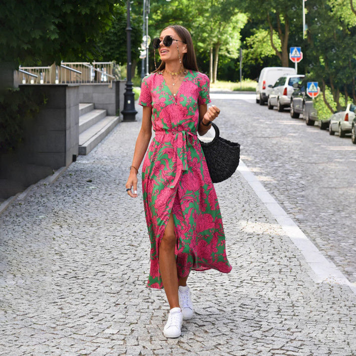 Vrouw in roze-groene bloemenjurk loopt op geplaveide straat, draagt zonnebril en witte sneakers. Zomermode, stedelijke stijl, casual outfit.