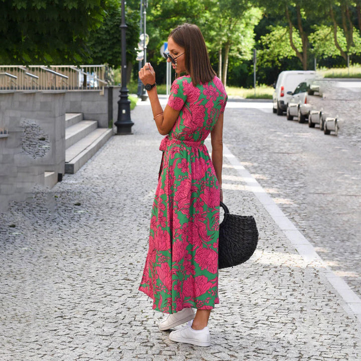 Vrouw in roze-groene bloemenjurk op geplaveide straat, draagt zonnebril en zwarte tas. Zomermode, casual stijl, stedelijke achtergrond.