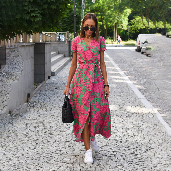 Vrouw in roze-groene bloemenjurk loopt op geplaveide straat, draagt zwarte tas en witte sneakers. Zomermode, stedelijke stijl, casual outfit.